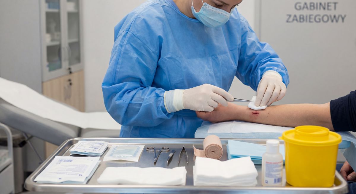 Pielęgniarka w niebieskim fartuchu i maseczce opatruje ranę na przedramieniu pacjenta w gabinecie zabiegowym, korzystając z jałowych gazików i narzędzi ułożonych na tacy, co ilustruje kompetencje specjalisty w pielęgniarstwie chirurgicznym oraz dbałość o bezpieczeństwo pacjenta.