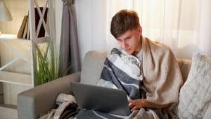 Mężczyzna owinięty kocami korzysta z laptopa na kanapie w domu.