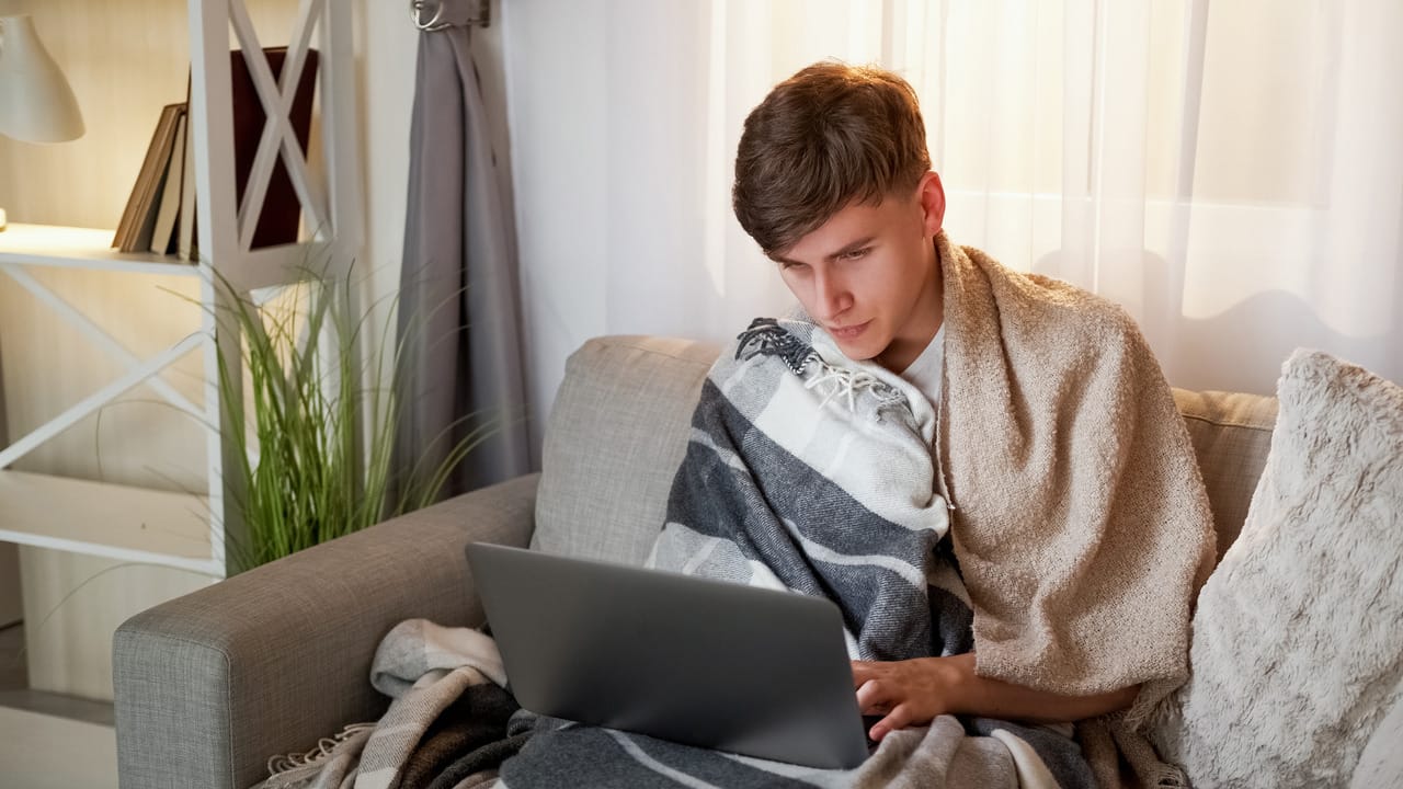 Mężczyzna owinięty kocami korzysta z laptopa na kanapie w domu.